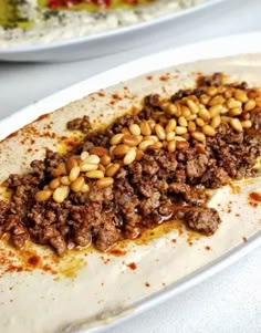 a burrito covered in ground beef and pine nuts on a white plate with other plates behind it