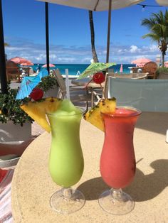 two drinks sitting on top of a table next to each other under an umbrella near the ocean