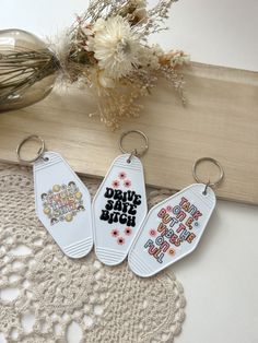 three keychains with different designs on them sitting next to a lace doily