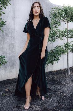 a woman standing in front of a concrete wall wearing a green wrap dress and sandals