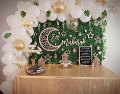 a table topped with balloons and decorations next to a wall covered in greenery that says eid mubara