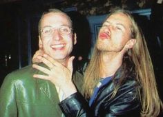 two men standing next to each other in front of a building and one man has long hair