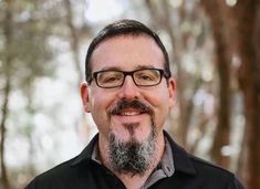 a man wearing glasses and a black shirt smiles at the camera with trees in the background