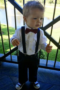 a little boy wearing a bow tie and suspenders standing in front of a gate