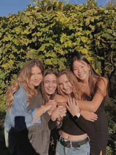 four girls are posing together in front of some bushes and trees with their arms around each other