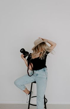 a woman sitting on a stool holding a camera and taking pictures with her hat over her head