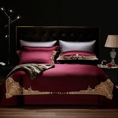 a bed with red sheets and gold trimmings in a dark room, next to a lamp