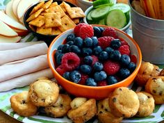 an assortment of fruits and crackers on a plate next to dips, cucumber wedges, apple slices, and other snacks