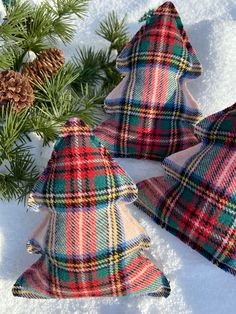 three plaid christmas tree ornaments sitting in the snow next to pine cones and evergreen branches