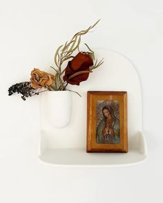 a white shelf with a painting and flowers in it on top of the shelf is a small wooden frame