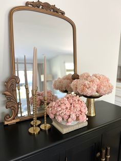 a black dresser topped with pink flowers and gold candles next to a large mirror on top of it