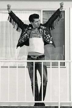 a man standing on a balcony with his arms in the air and wearing a leather jacket