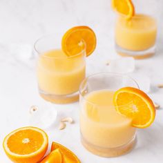 two glasses filled with orange juice on top of a white counter next to sliced oranges