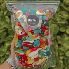 a bag filled with lots of different colored candies next to a bush full of green leaves