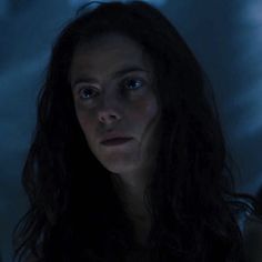 a woman with long dark hair and blue eyes stares into the distance in a dimly lit room