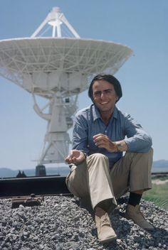 a man sitting on the ground in front of a radio telescope with his hand out