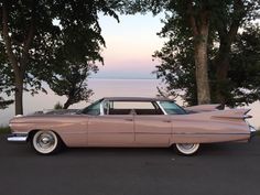 an old pink car is parked in front of some trees and the water behind it