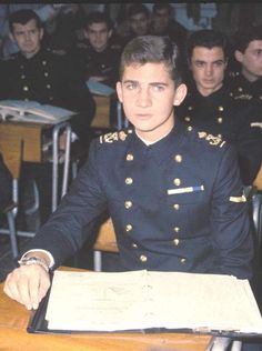 a man in uniform sitting at a desk with other men behind him and writing on a piece of paper