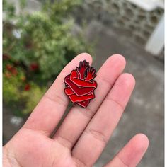 a red heart shaped pin sitting on top of a person's hand in front of some flowers