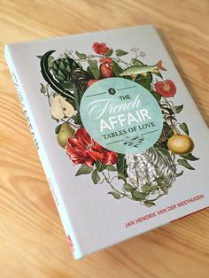 the front cover of a book on a wooden table with flowers and fruit around it