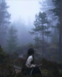 a woman sitting in the middle of a forest on a foggy day with trees