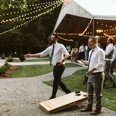 two men in ties are playing cornhole game at an outdoor wedding reception with string lights