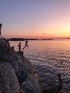 some kids are playing on the rocks by the water at sunset or sunrise, while others swim in the lake