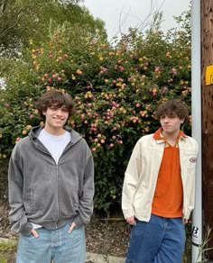 two young men standing next to each other in front of a bush with pink flowers