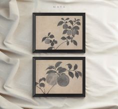 two framed pictures with leaves on them sitting on top of a white bed sheet covered in sheets