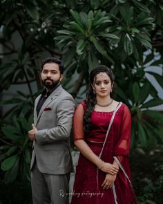 a man and woman standing next to each other in front of some green plants with trees behind them