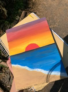 a hand holding a spiral notebook with a sunset painting on it and the ocean in the background