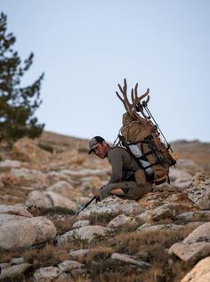 a man kneeling on top of a rocky hillside next to a backpack filled with antlers