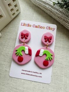 pink and green handmade earrings with bows on them sitting on a white card next to a plant