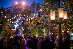 people are gathered at an outdoor event in the evening with lights strung from trees and lanterns