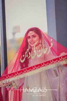 a woman in a white dress and veil holding up a red shawl with arabic writing on it