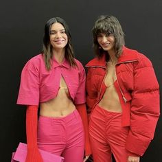 two women standing next to each other wearing red outfits and holding pink bags in front of a black wall