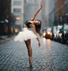 a ballerina is dancing in the rain on a cobblestone street with cars behind her