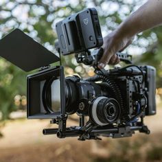 a camera being used to film an outdoor scene with trees in the background and someone's hand reaching for it