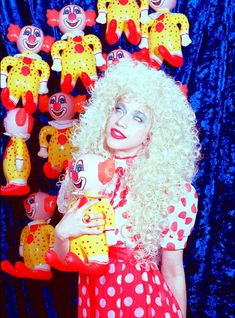 a woman with blonde hair holding a teddy bear in front of many clowns hanging on the wall