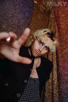 a man with blonde hair and blue eyes posing in front of a gold wall holding his hand up