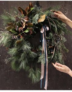 two hands are holding a wreath with pine cones and evergreens on it, while another hand holds a ribbon around the wreath