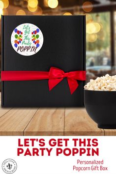 a black box with red ribbon and popcorn in it on a wooden table next to a bowl of popcorn