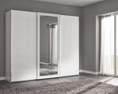 a white wardrobe with mirrored doors in a room next to a window and rug on the floor