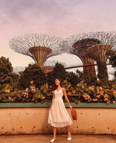 a woman in a white dress holding a brown purse and standing next to some trees