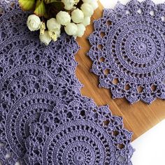 two crocheted doily sitting on top of a wooden cutting board next to flowers