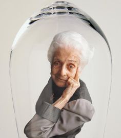 an elderly woman in a glass case with her hand on her chin and looking at the camera