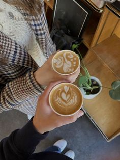 two people are holding cups with coffee art on the top and one is holding a plant