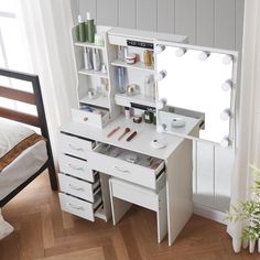 a white vanity table with drawers and lights on the mirror above it, in front of a window