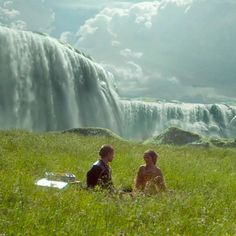 two people sitting in the grass near a waterfall