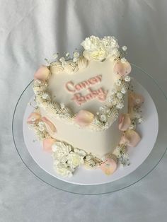 a heart shaped cake on a plate with white flowers and pink lettering that says cozy baby